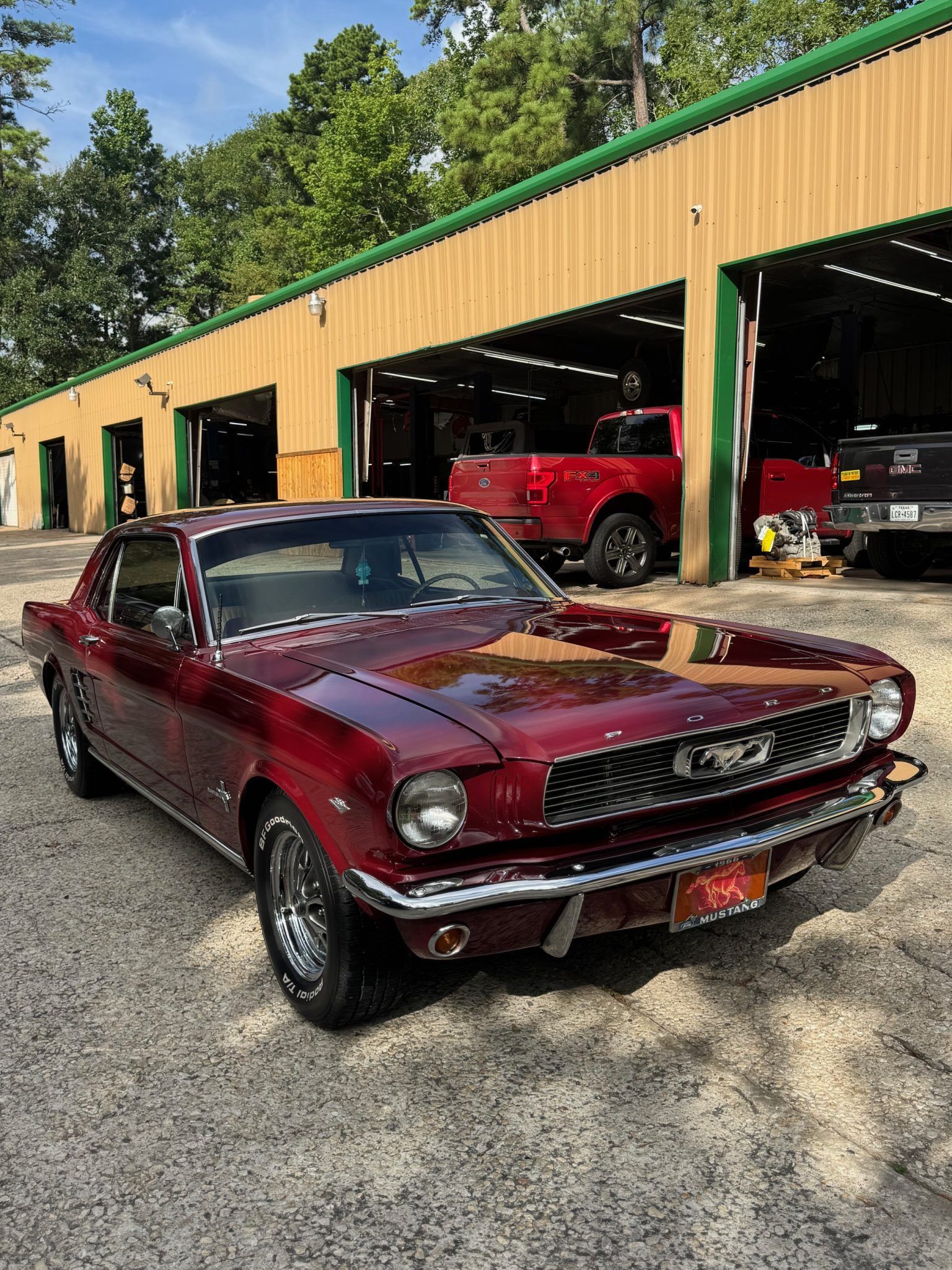 Maroon classic Mustang parked outside a garage with other cars inside and a wooded background