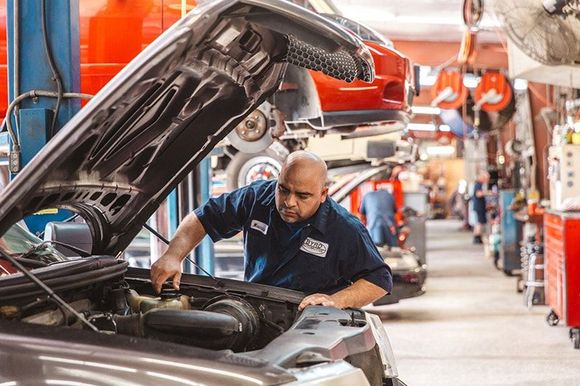 Mechanic inspecting a car with hood open in an auto repair shop, with tools and vehicles in the background