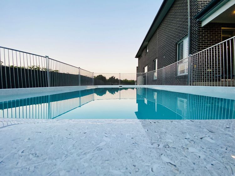 A large swimming pool is surrounded by a fence and a brick building.