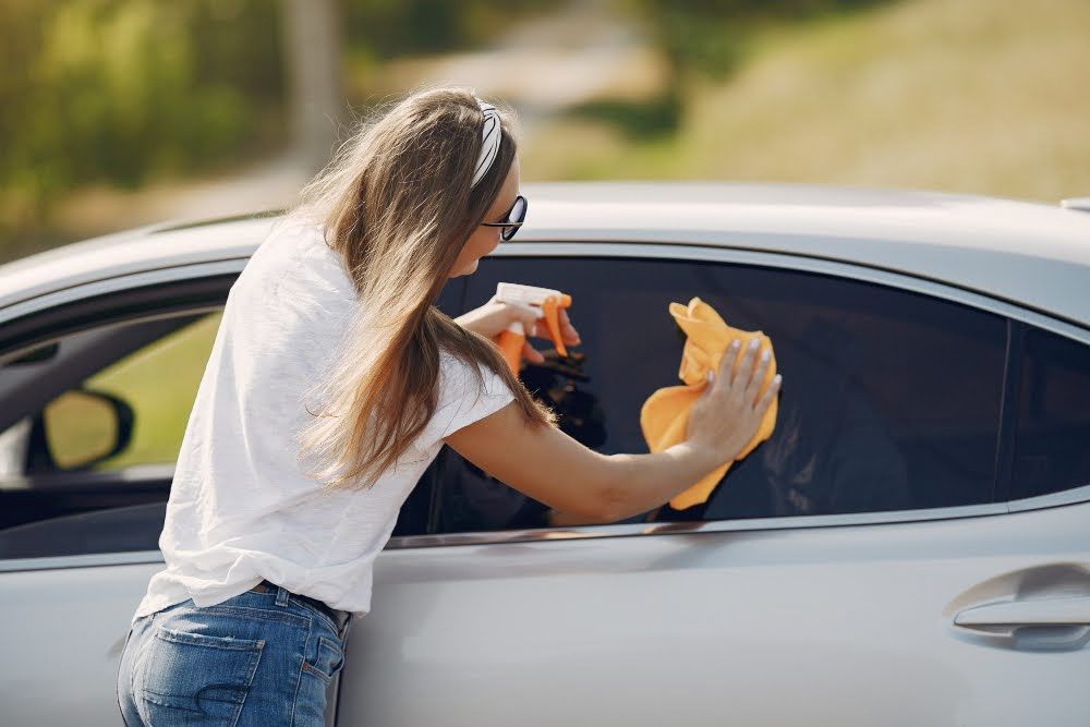 A woman wiping a tinted window of a car using a microfiber cloth.