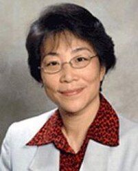 Studio headshot of a smiling person wearing glasses, a white blazer, and a red patterned shirt.