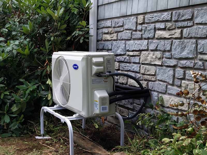 Outdoor air conditioner unit on a metal stand beside a stone wall and greenery