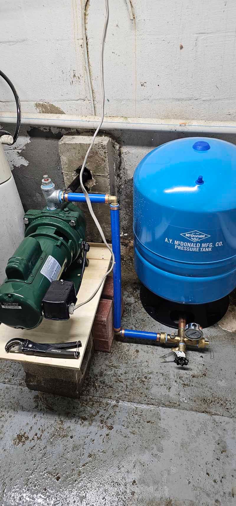 Green water pump beside blue pressure tank with PVC piping on a wet concrete floor