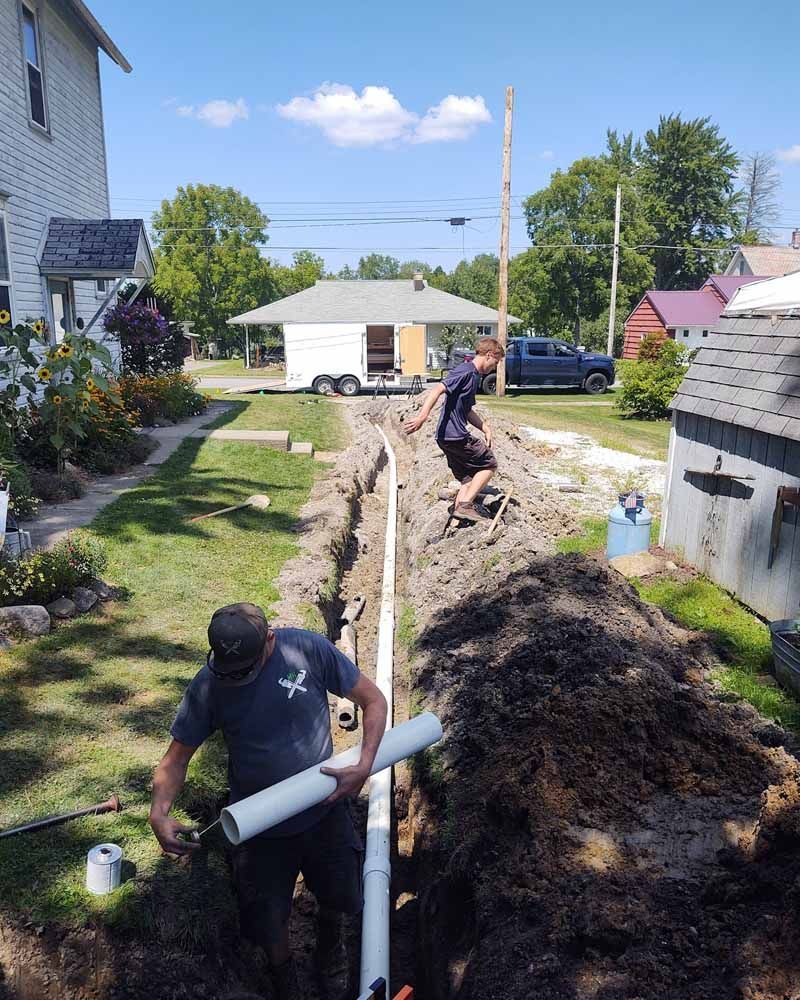Workers install white PVC piping in a trench beside a house in a residential yard.