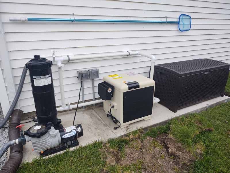 Pool equipment beside a house: pump, filter, heater, and large storage box along a white wall.