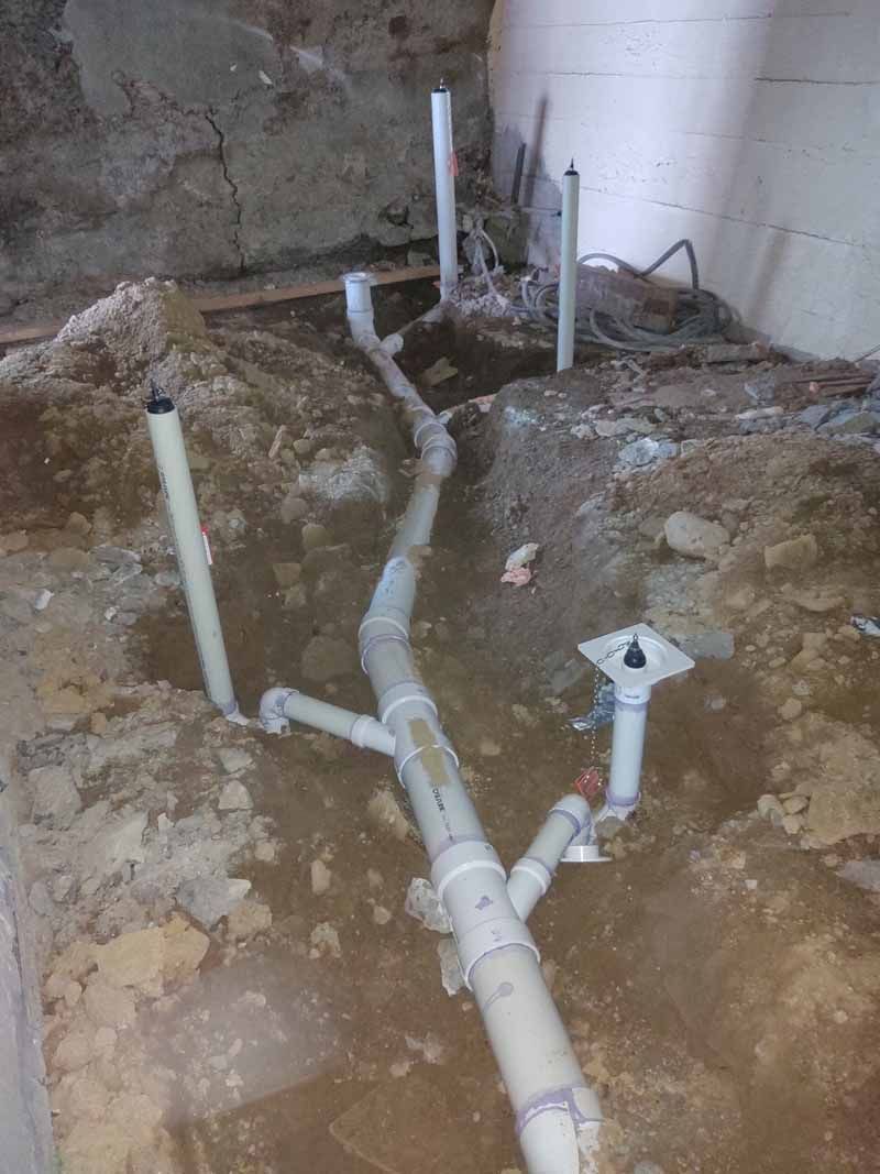 Exposed white plumbing pipes and drain lines in a rough, unfinished basement floor area