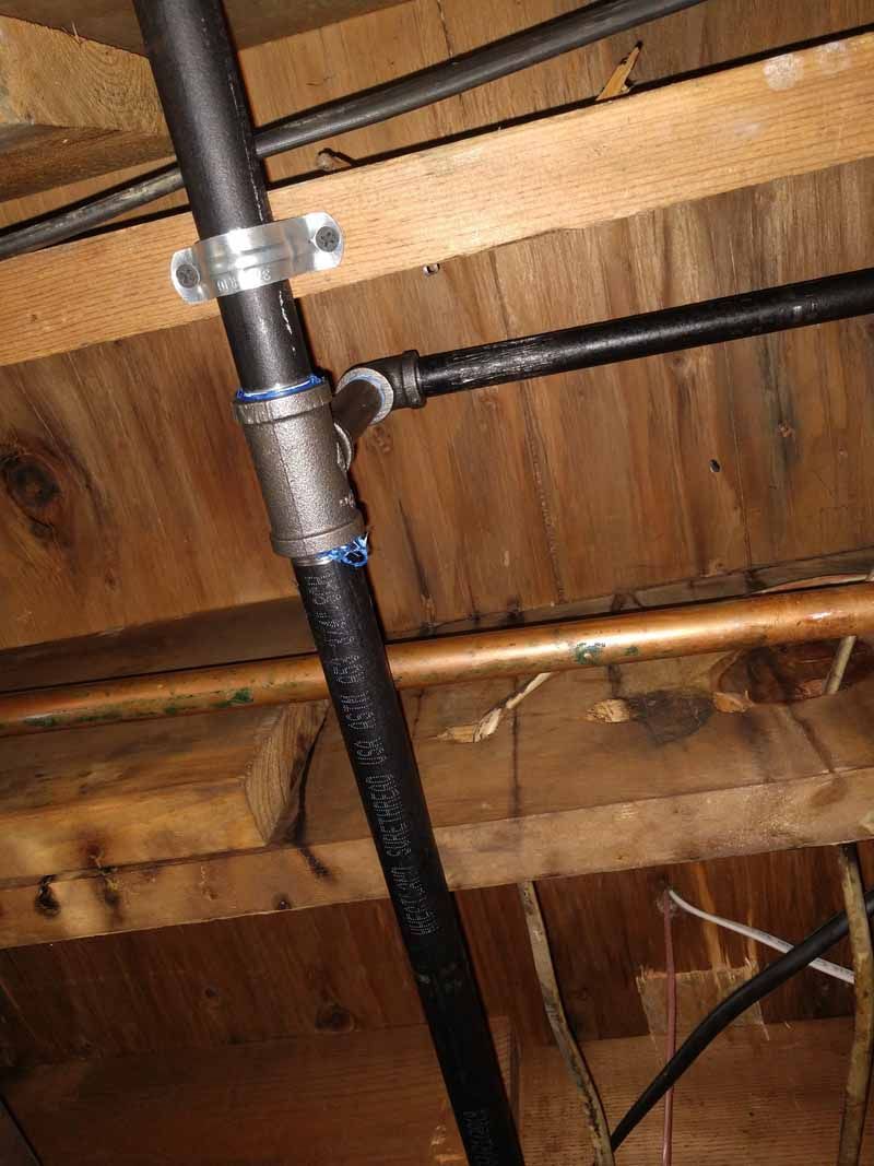 Black pipe with metal clamp mounted to wooden joists in a basement ceiling