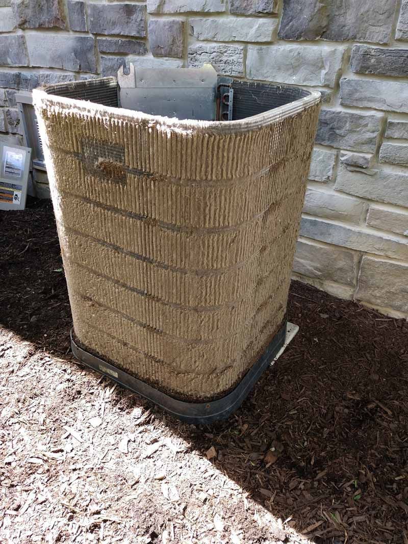 Outdoor woven air conditioner cover beside a stone wall on mulch.