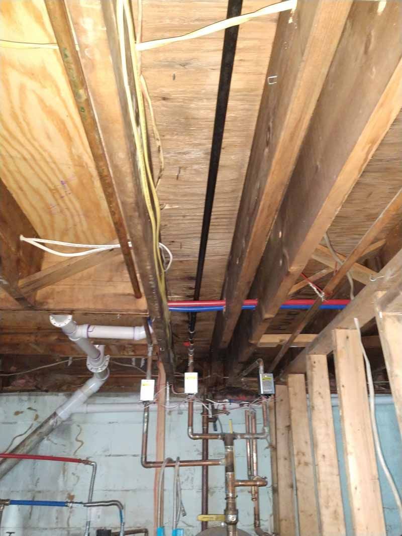 Unfinished basement ceiling with exposed wooden joists, pipes, and electrical wiring.