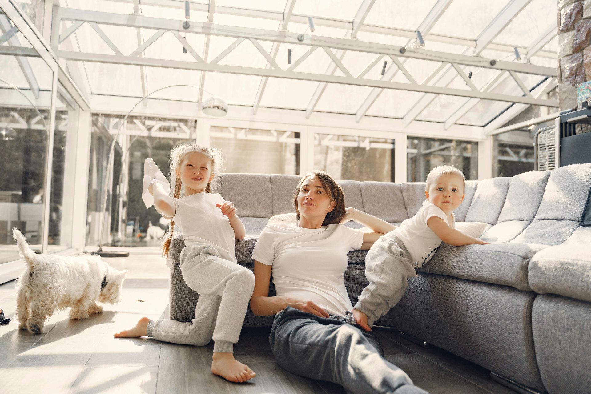 Woman with two children sitting on a gray couch in a bright living room, with a small white dog nearby.