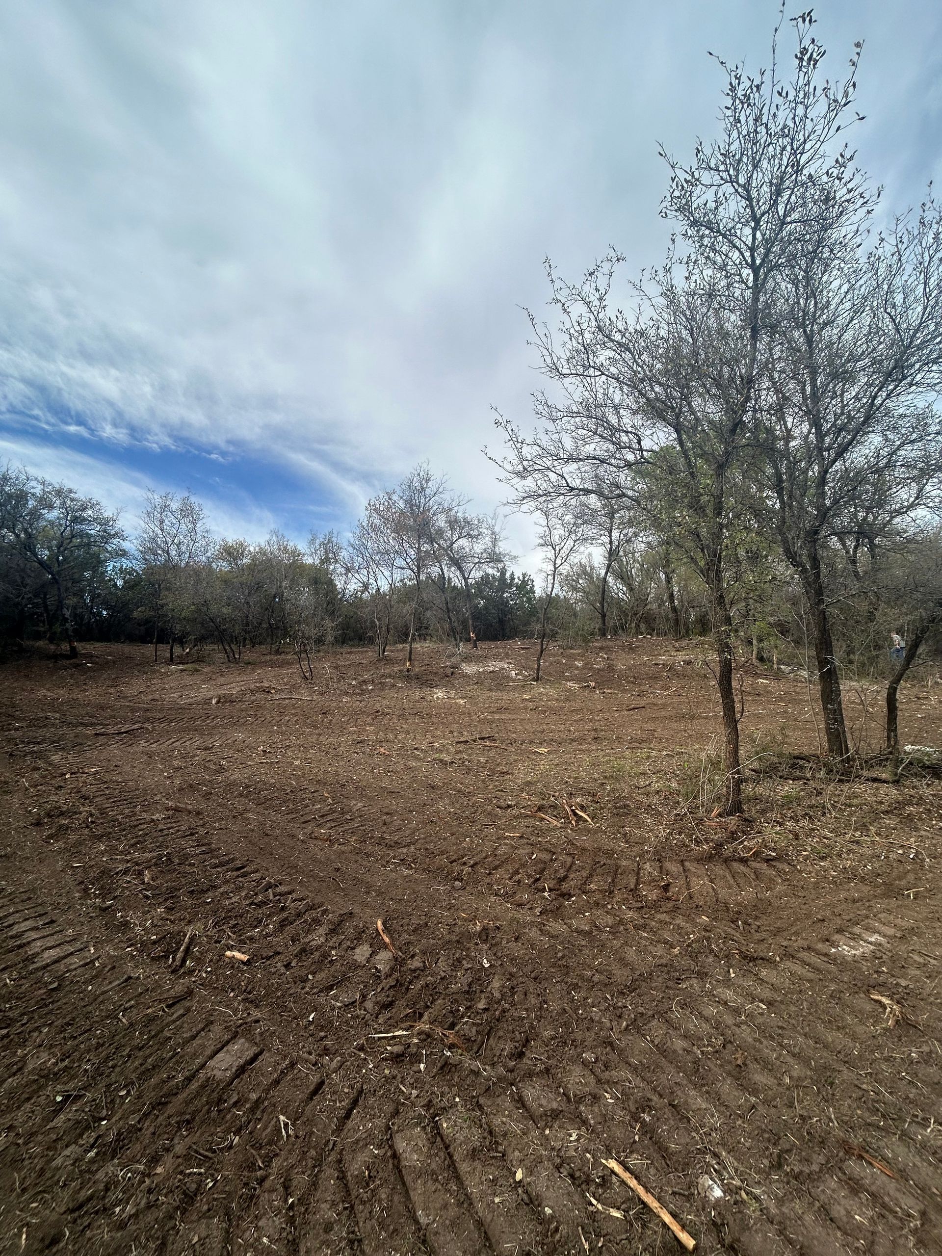 A field of freshly tilled brown soil sits beneath a cloudy blue sky, bordered by a line of trees in the background.