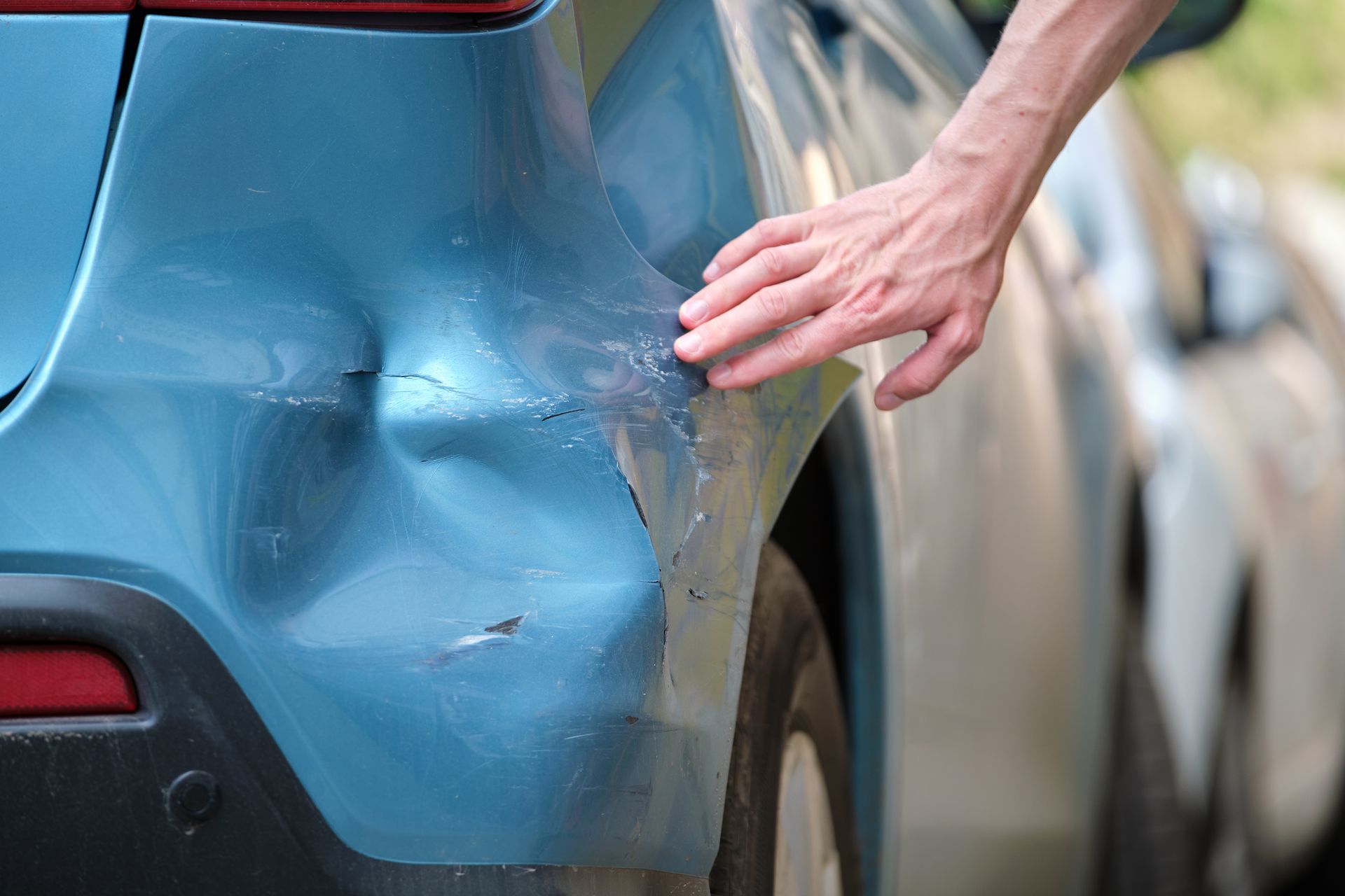 Person's hand touching a dented, scratched blue car bumper after a minor collision.