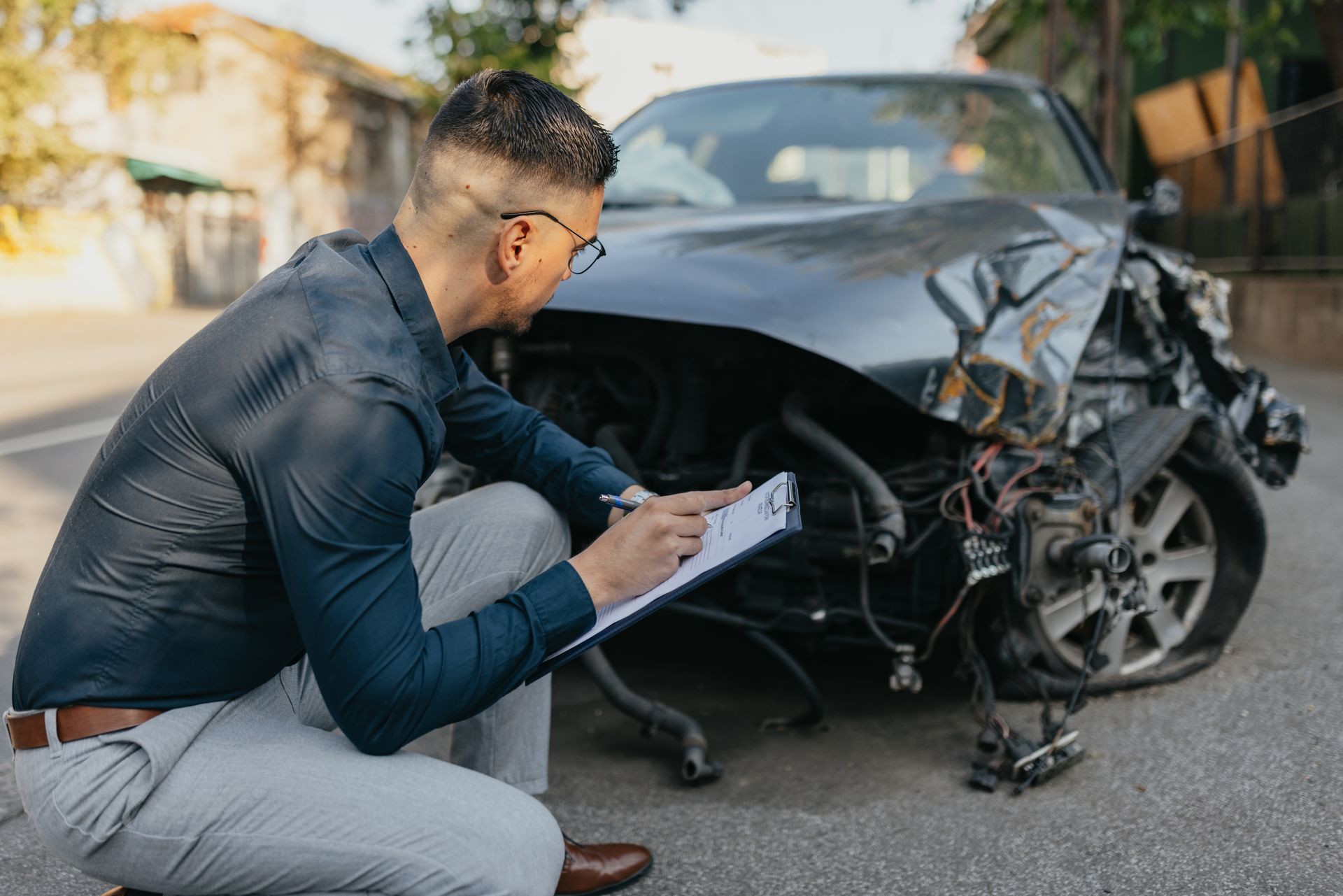 Man in a blue shirt inspects a heavily damaged car, writing on a clipboard outdoors.