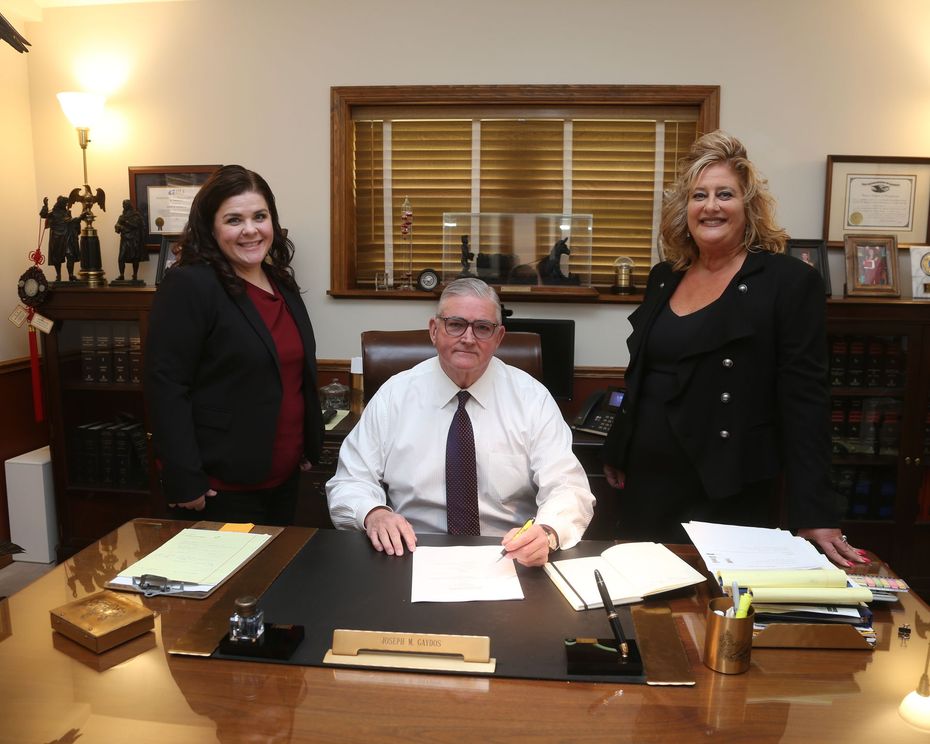 Three people at a desk: two women flanking a man sitting, all in business attire, in an office.