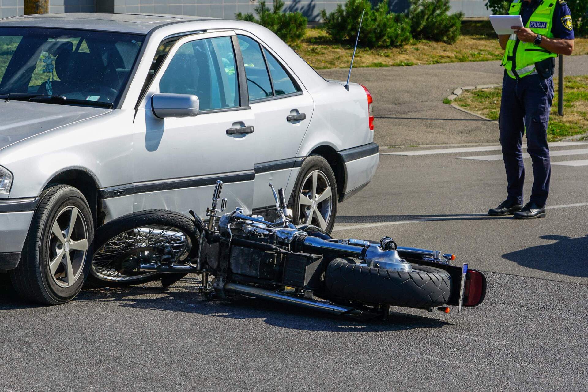A silver car has crashed into a motorcycle lying on its side in a street; a police officer stands nearby.