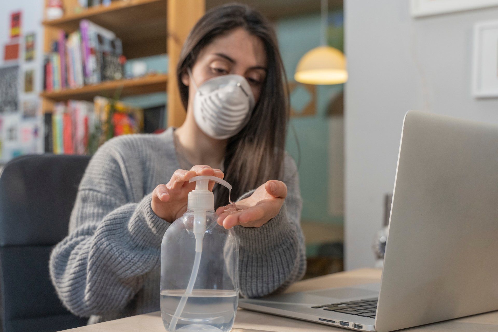 Woman wearing a mask, using hand sanitizer at a desk with a laptop. Bookshelf in the background.