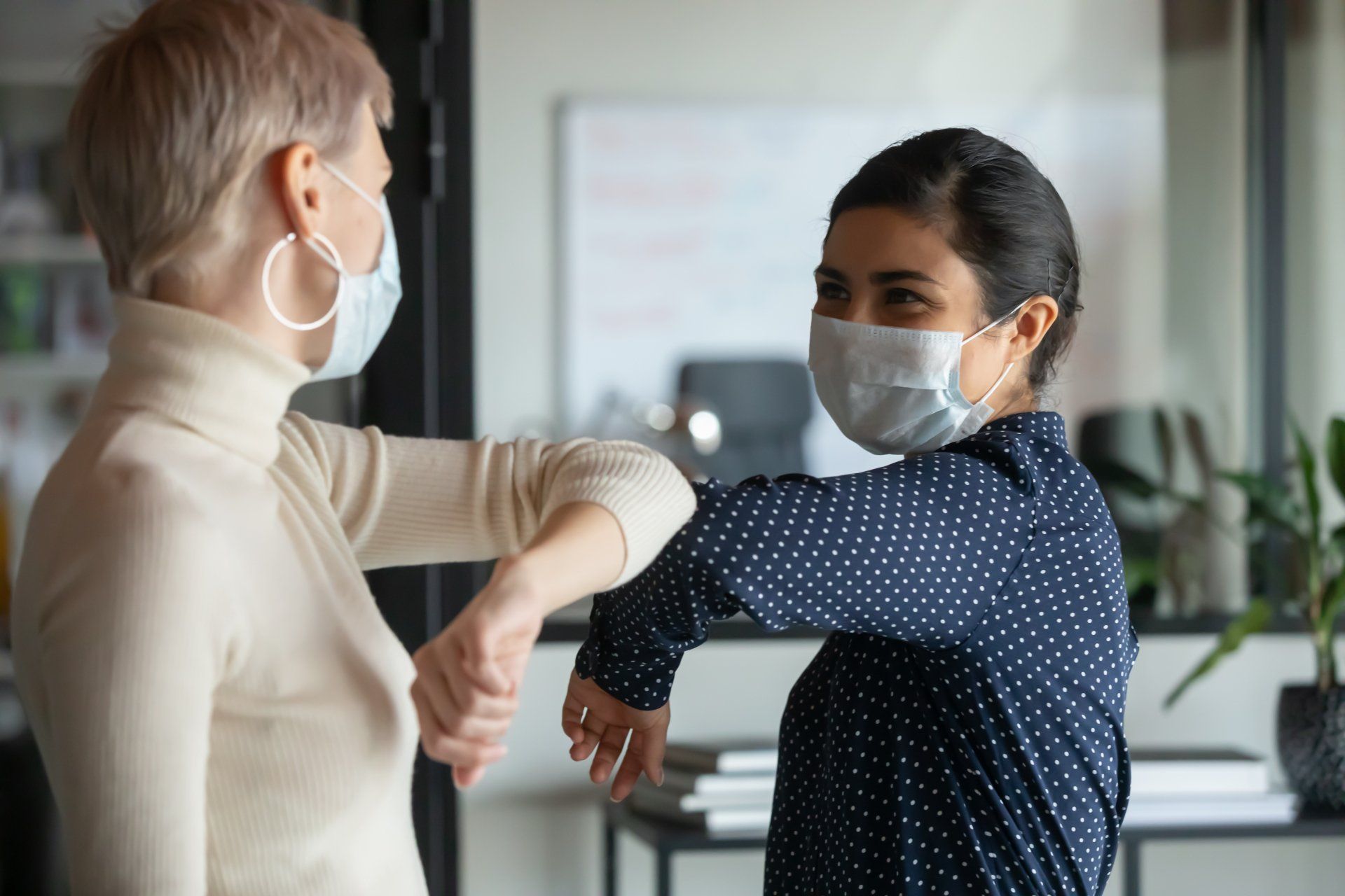Two women in masks bump elbows in an office; one smiles.