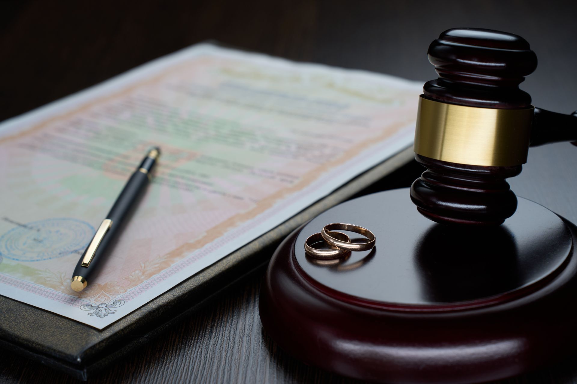 Gavel and wedding rings on a stand next to divorce papers and a pen.