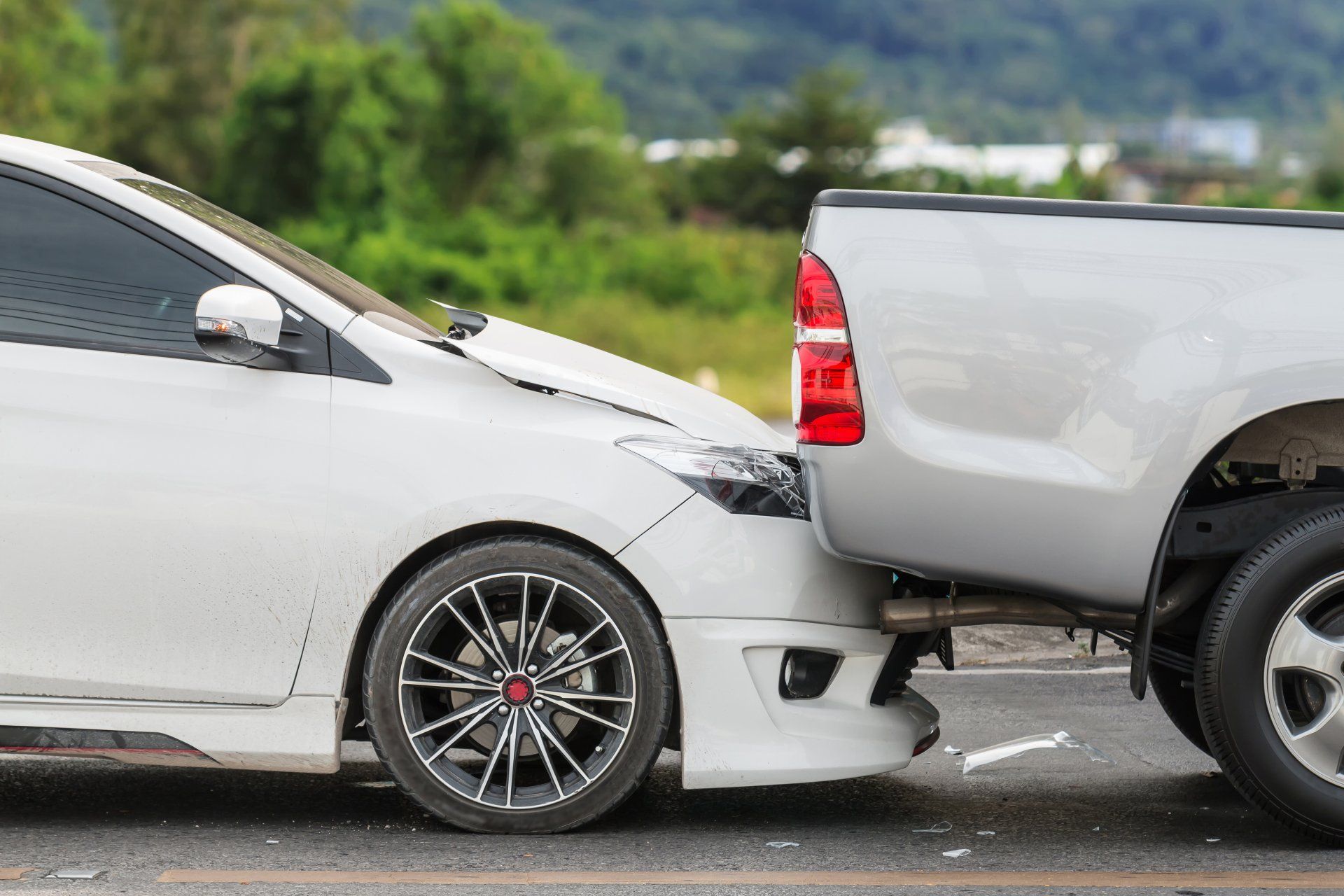 White car with front end damaged, rear-ended by silver pickup truck on a road.