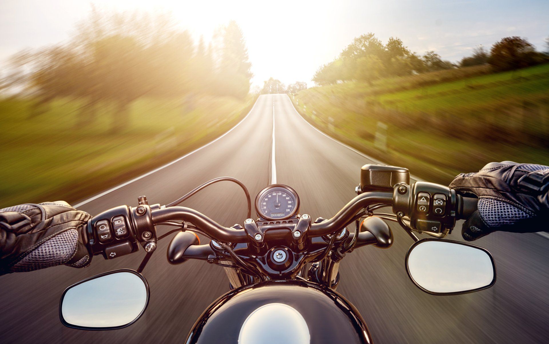 Motorcycle handlebars and rider's view of open road ahead, blurred background with bright sun.