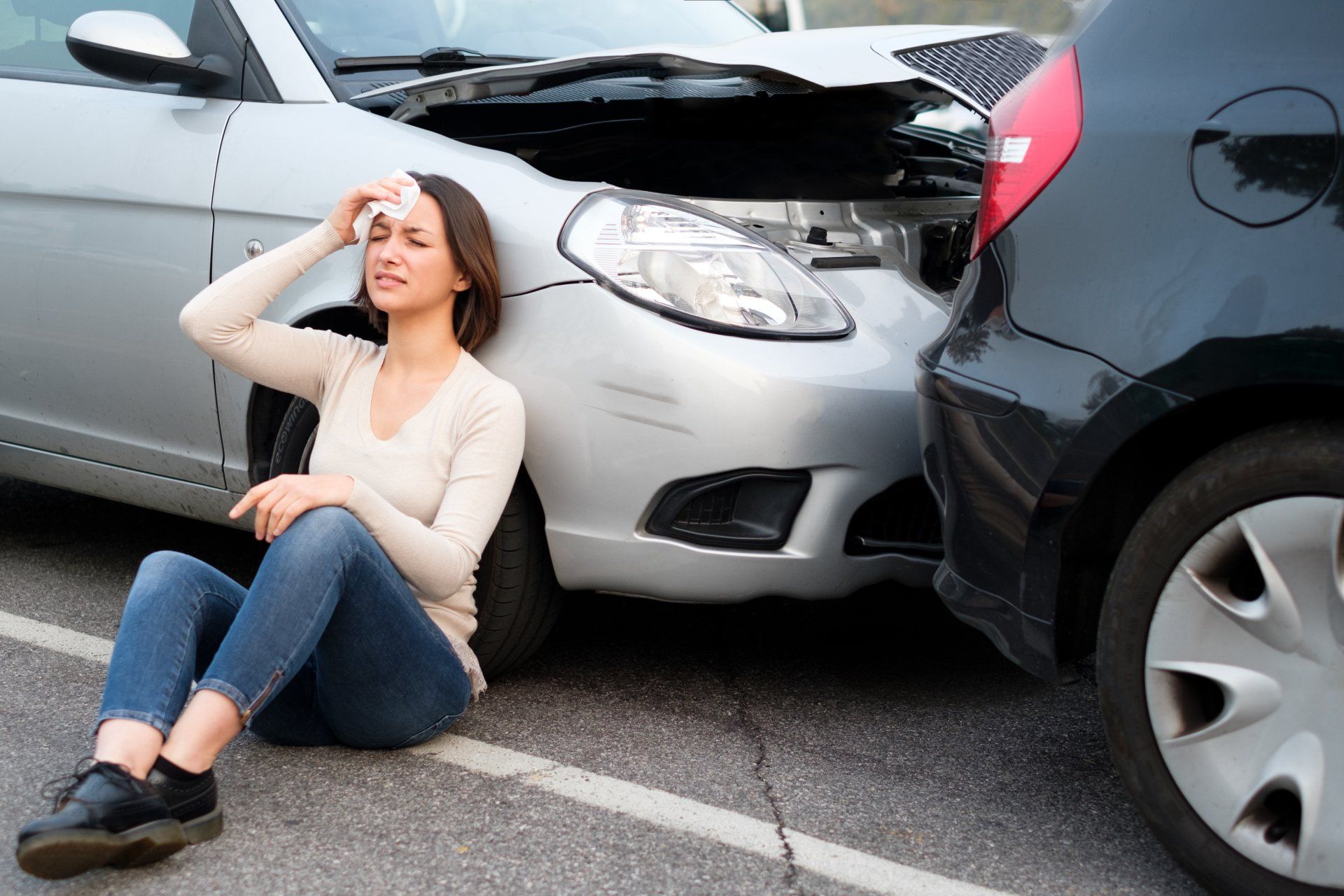 Woman injured at a car accident scene, sitting near damaged vehicles. Silver car front and black car rear.
