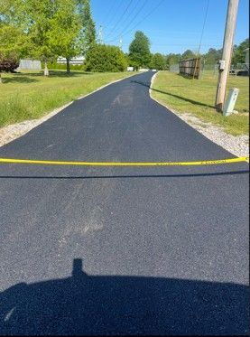 Newly paved asphalt road with yellow caution tape, flanked by green grass and trees under a clear sky.