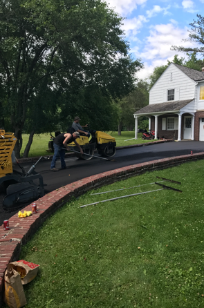 Asphalt driveway paving in progress. Workers operating a machine, green grass, brick border, white house, trees.