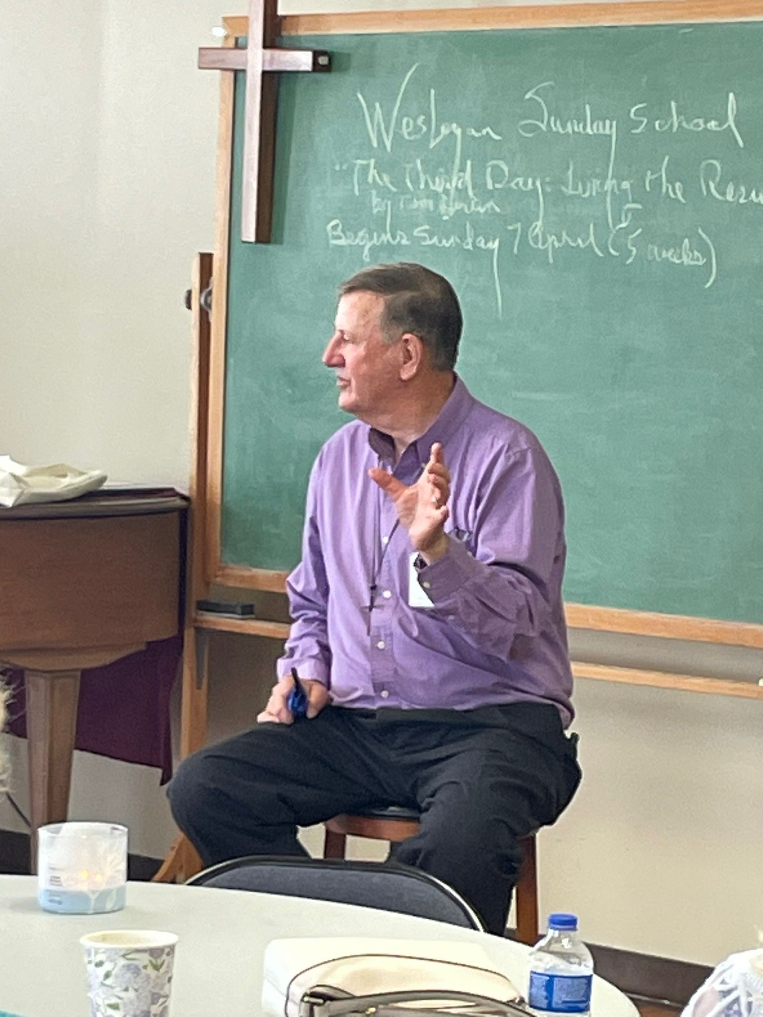 Man in purple shirt speaks, gesturing, in front of a chalkboard, cross on wall.