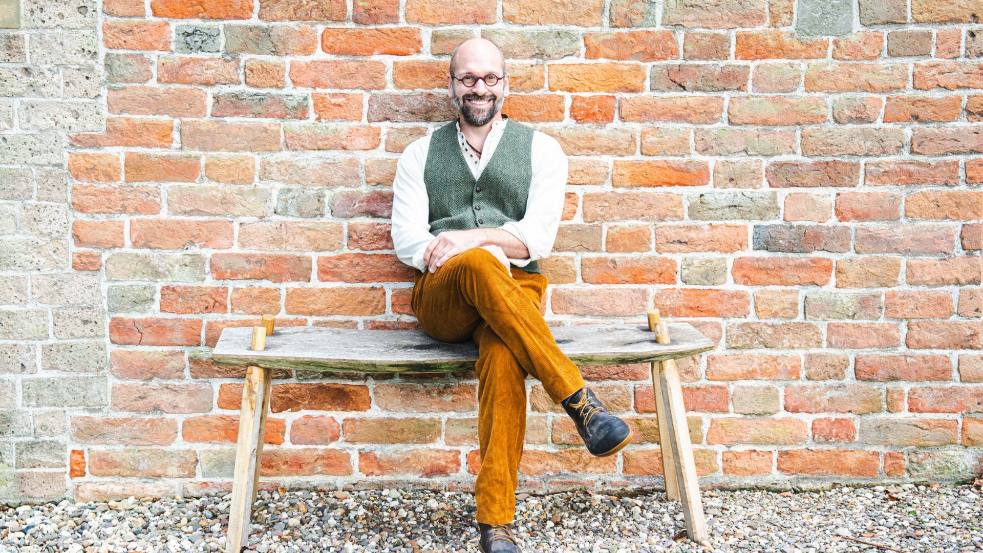 Bald man with glasses sits on wooden bench, legs crossed, against a brick wall.