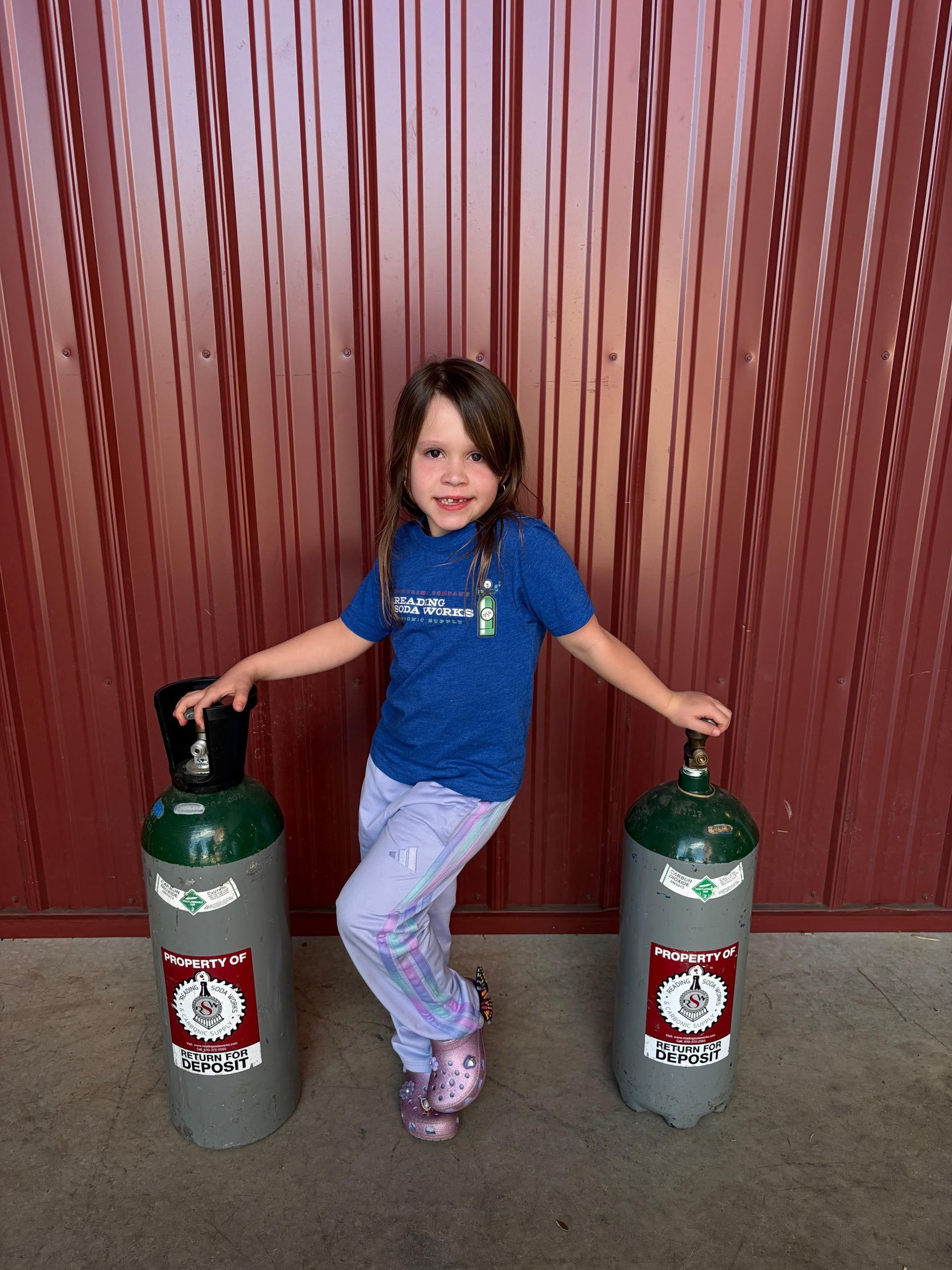 Child standing between two gas cylinders, holding one in each hand, in front of a red wall.