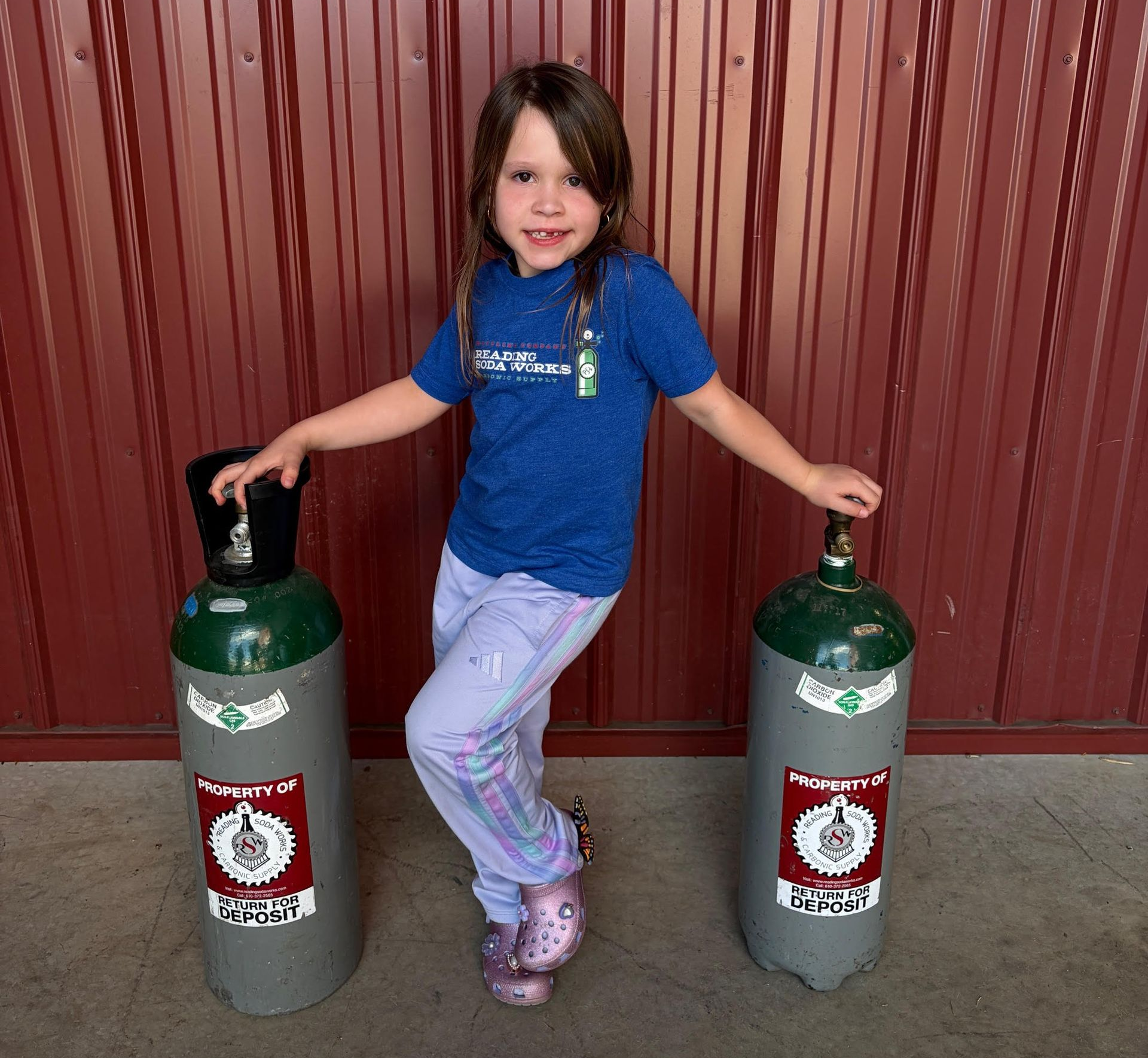 Child standing between two gas cylinders, wearing a blue shirt and purple boots, in front of a red wall