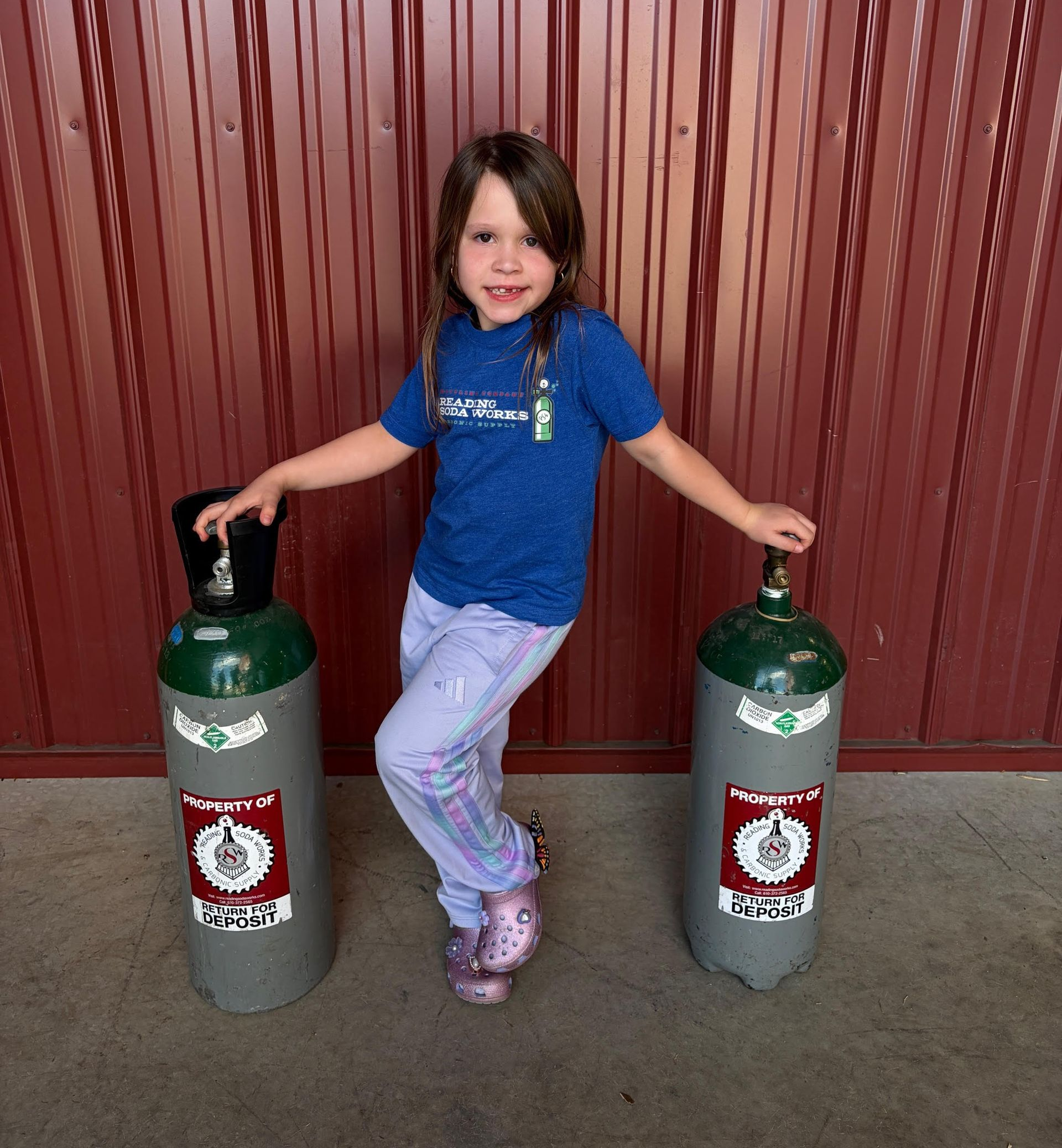 Child in a blue shirt standing between two green gas cylinders, smiling in front of a red wall.
