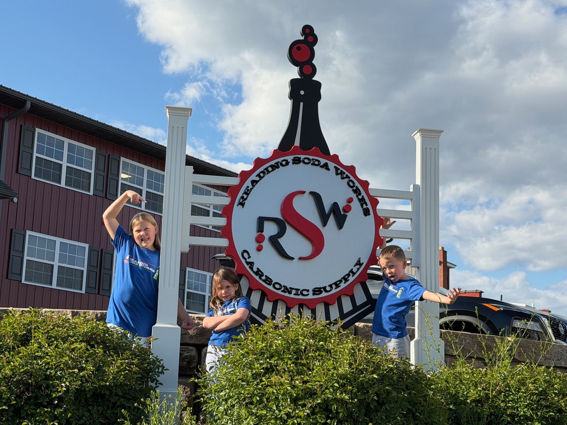 Children posing beside a large railroad crossing sign and hedge in front of buildings