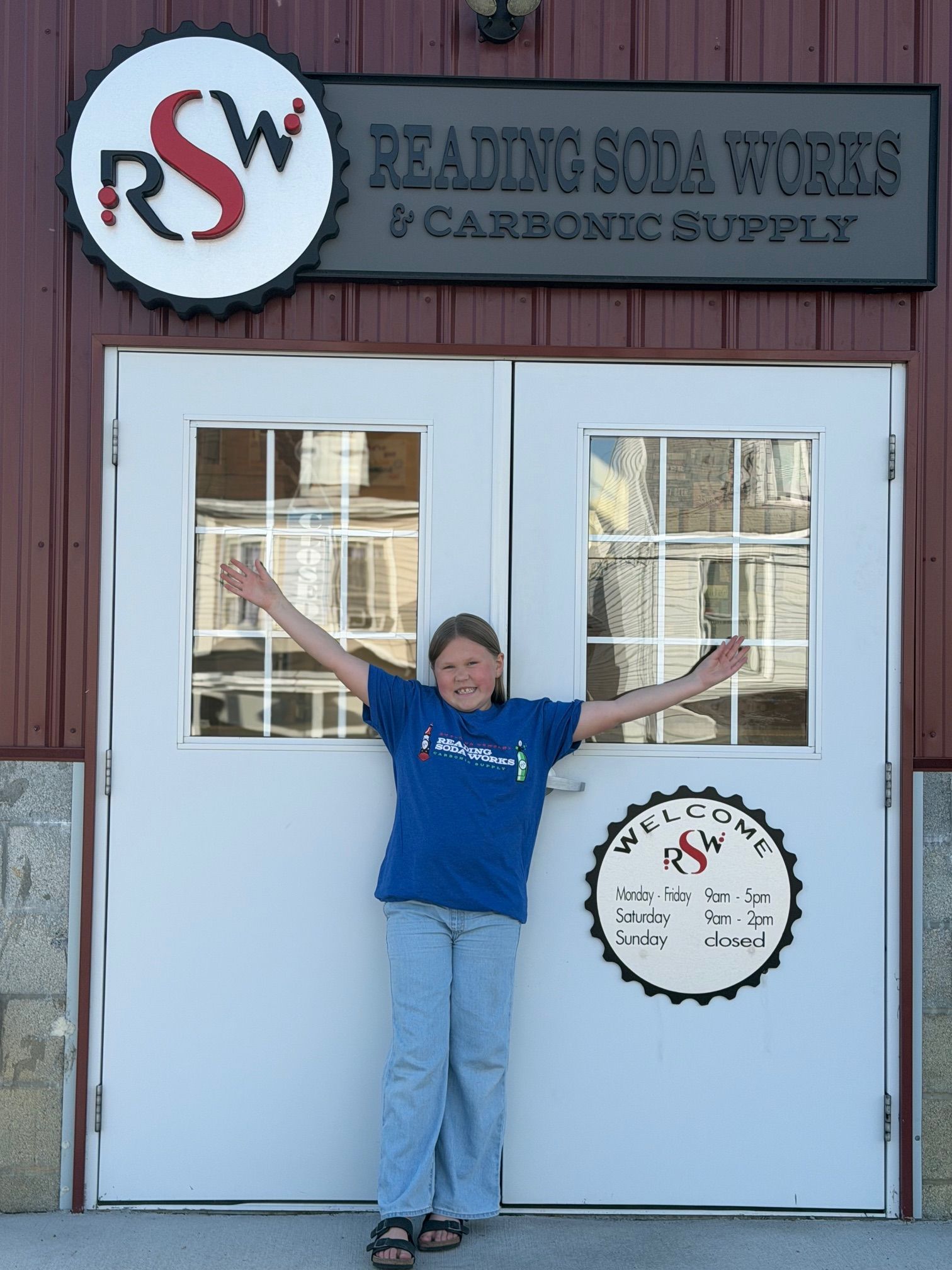 Woman in blue shirt standing with arms outstretched in front of Reading Soda Works storefront