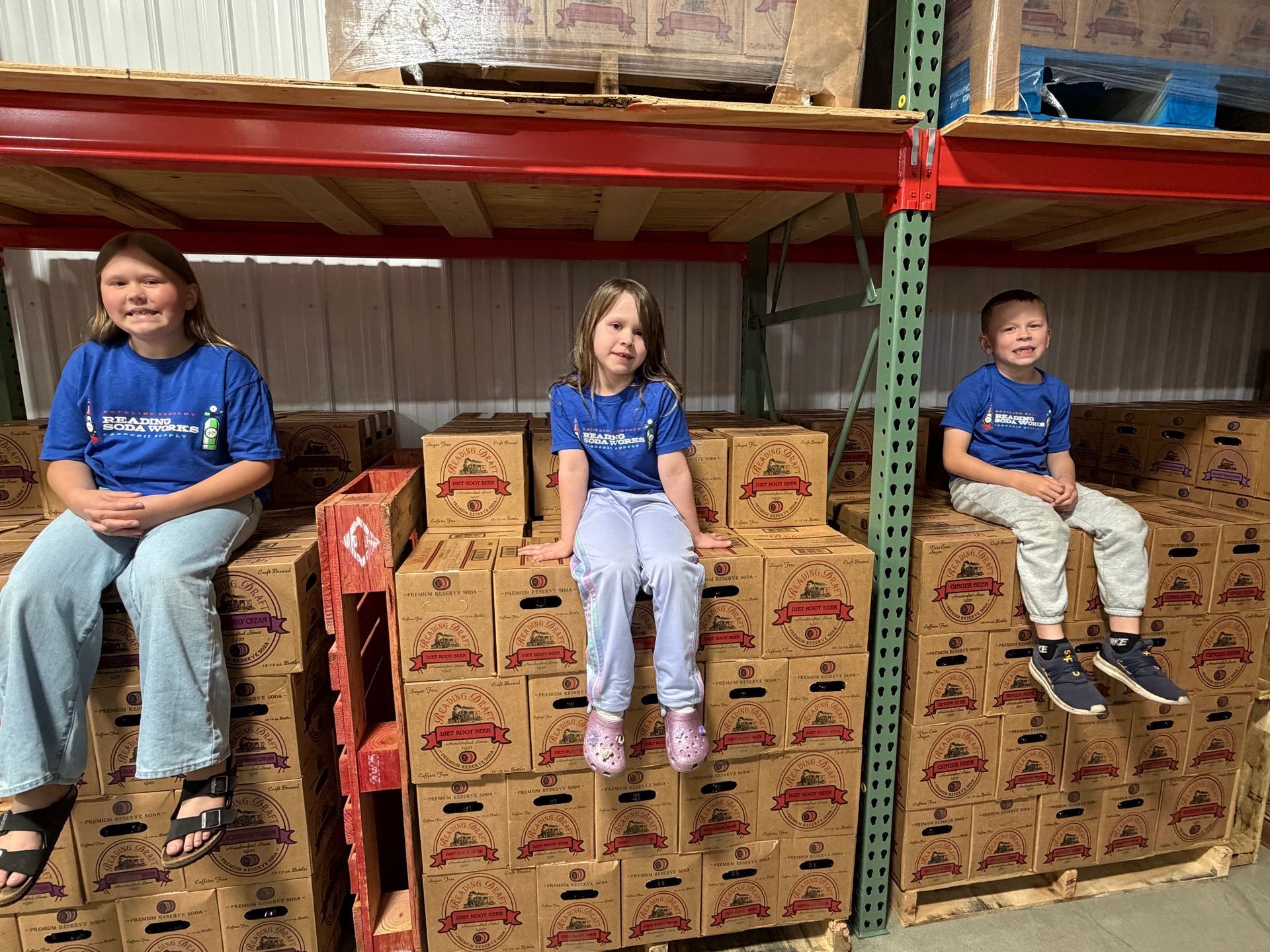 Three children in blue shirts sit on stacks of produce boxes in a warehouse aisle.