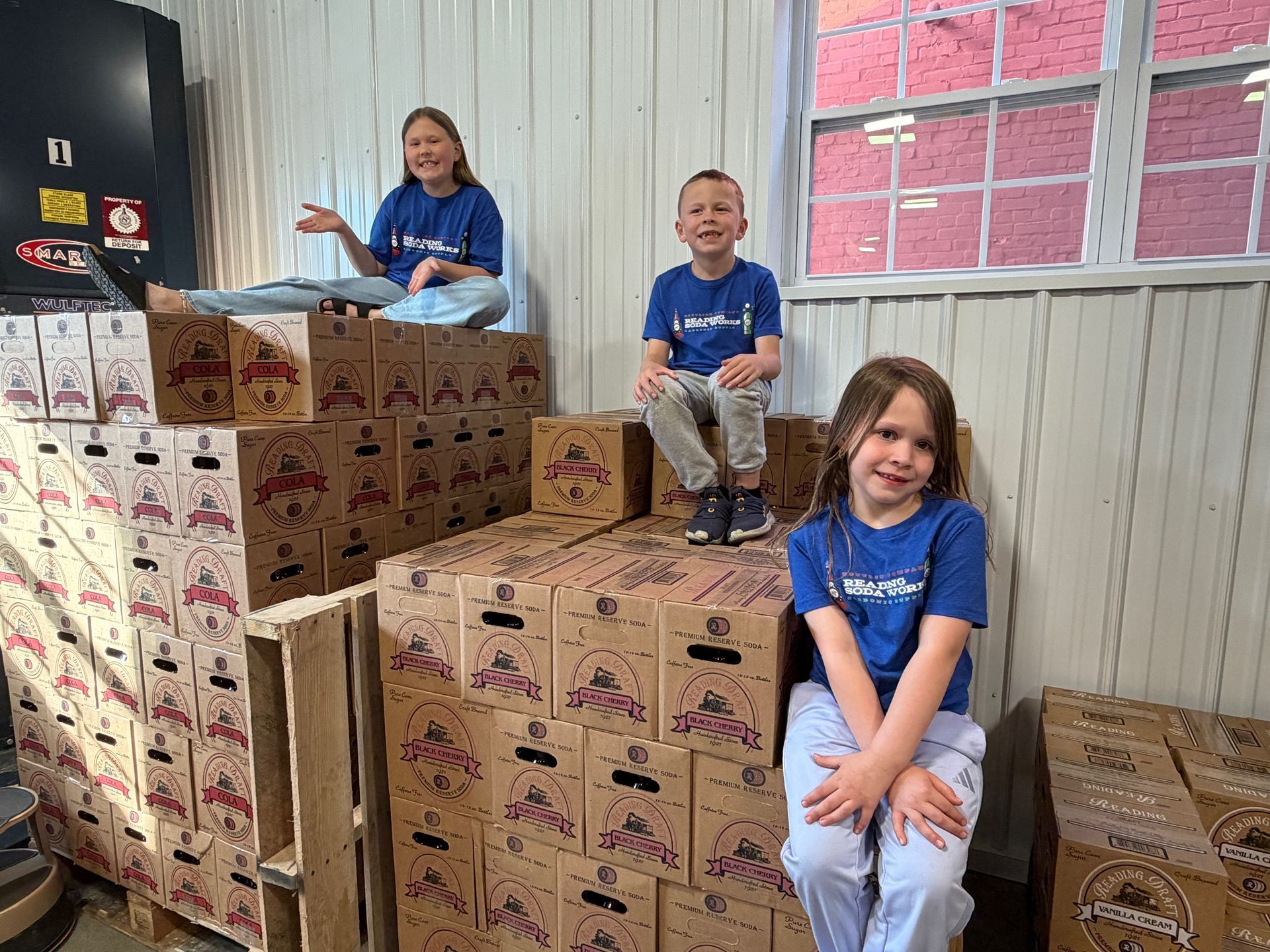 Children in blue shirts sitting on stacked cardboard boxes in a warehouse-like room