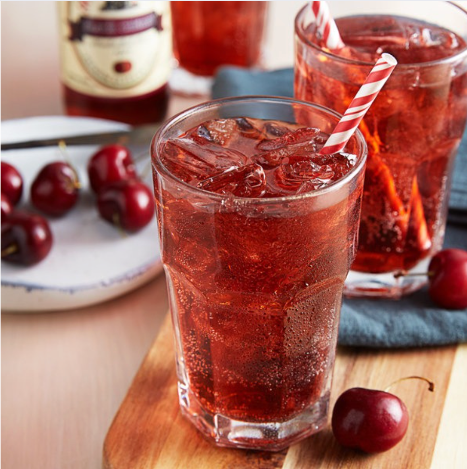 Two glasses of iced red cherry drink with striped straws on a wooden tray, cherries nearby
