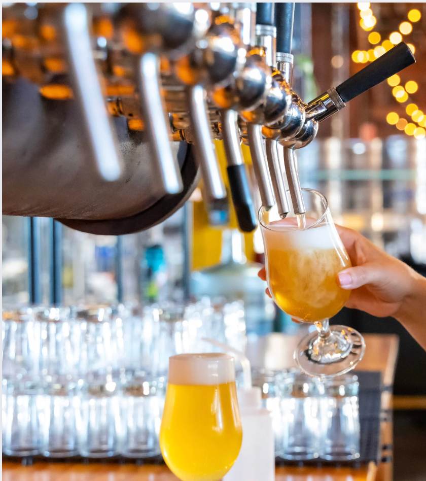 Hand pouring a beer from a tap into a stemmed glass in a bar, with other beer glasses nearby