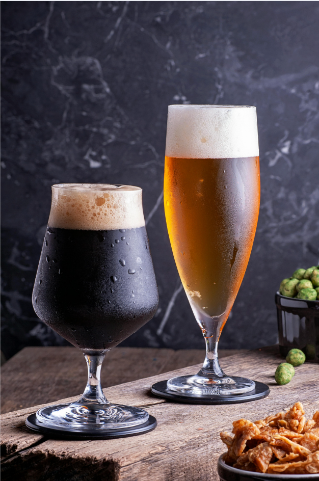 Two beer glasses on a wooden table, one dark stout and one amber beer, with hops and snacks nearby