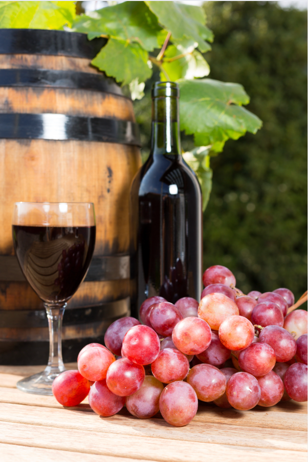 Wine bottle, glass, and red grapes beside a wooden barrel on a rustic table.