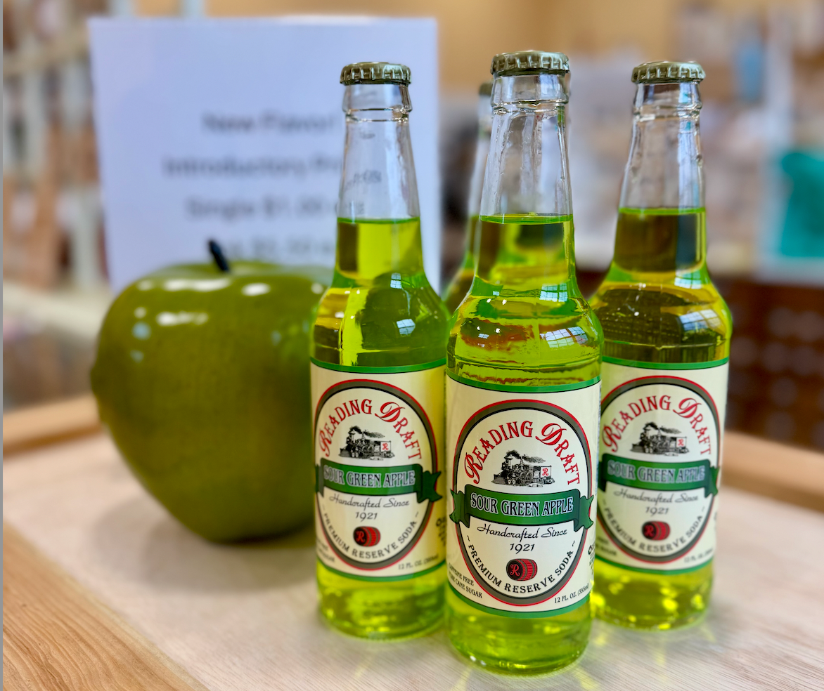 Three green glass bottles and a green apple-shaped container on a table, with a blurred sign behind them