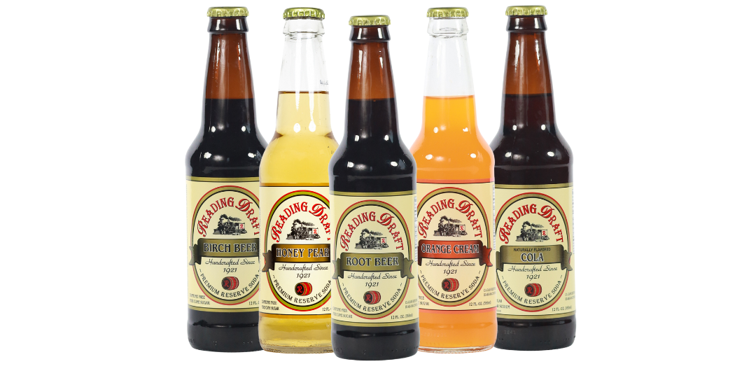 Five assorted glass soda bottles with vintage labels and dark, yellow, and orange drinks