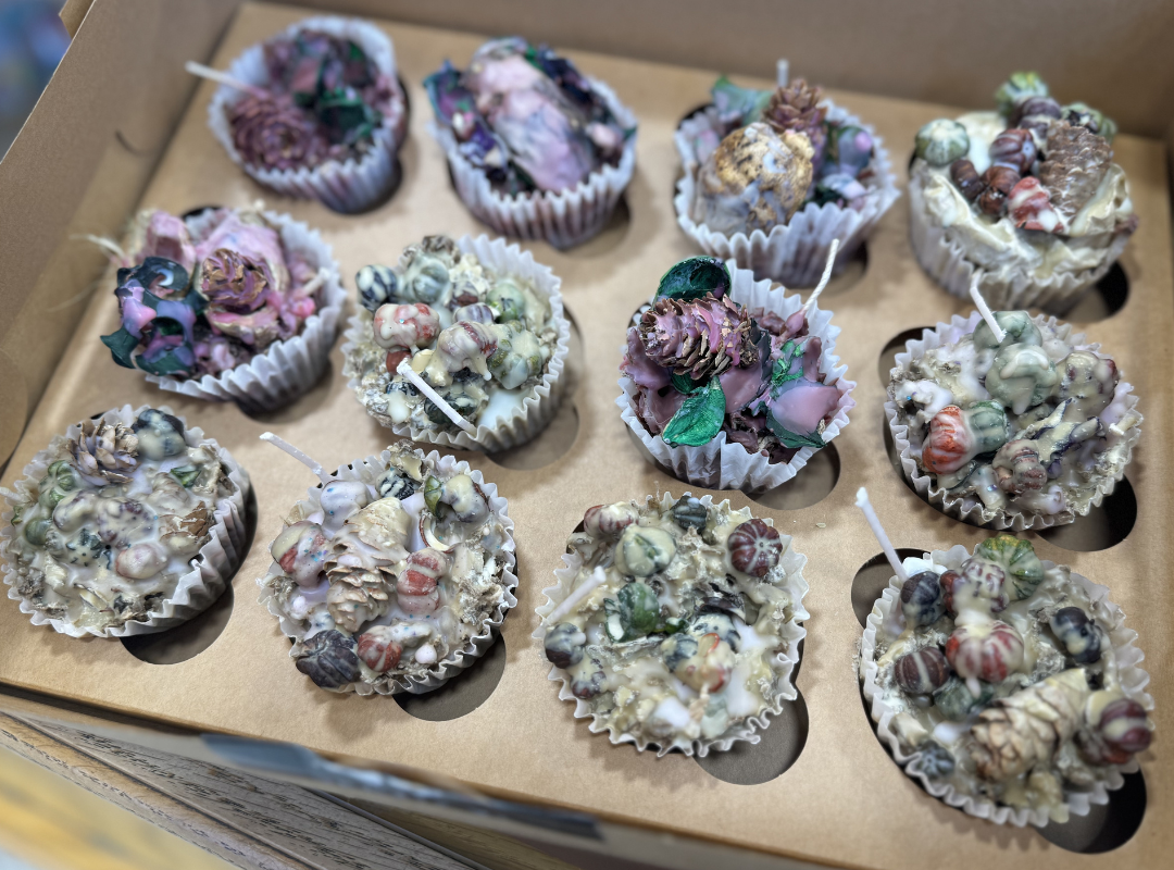 Box of frosted cupcakes with pastel pink and purple icing, sprinkled with bits, in a cardboard tray