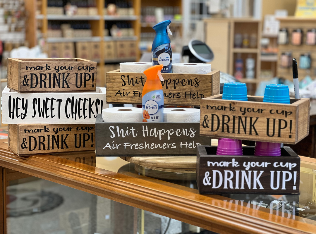 Rustic wooden signs with humorous phrases in a store display, including “Drink Up!” and “Shit Happens”