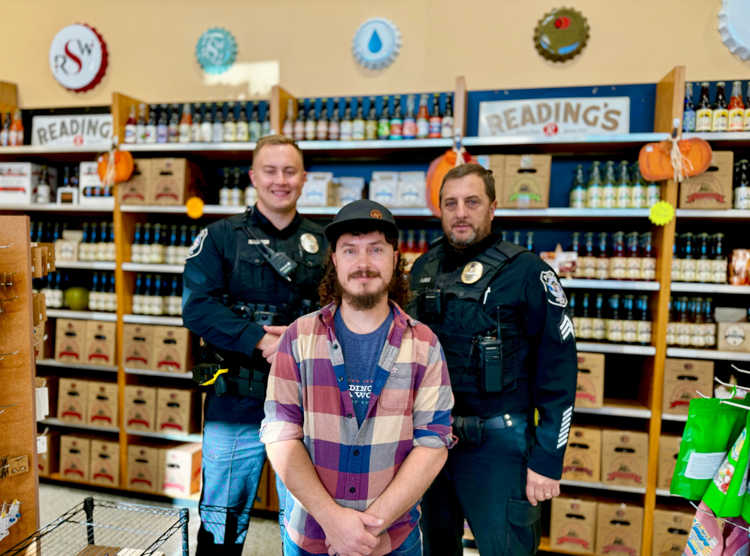 Three people pose in a store aisle with shelves of colorful jars and signs reading “READINGS.”