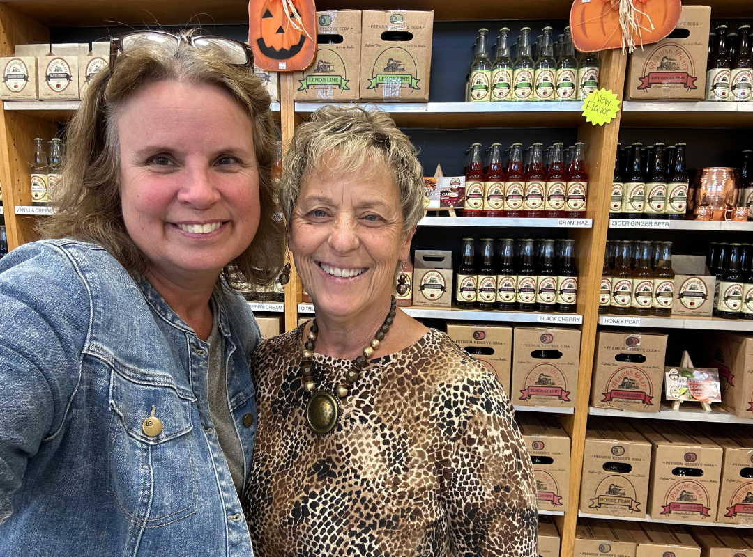 Two women smiling in a shop aisle with shelves of jars and fall decorations behind them