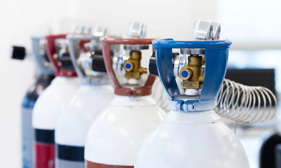 Row of gas cylinders with colored valve caps, lined up in a workshop or industrial setting.