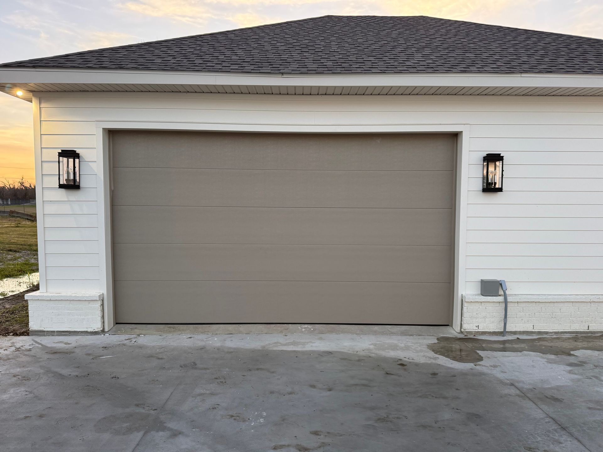 Tan garage door on a white building, flanked by black lights. Cement driveway with overcast sky.