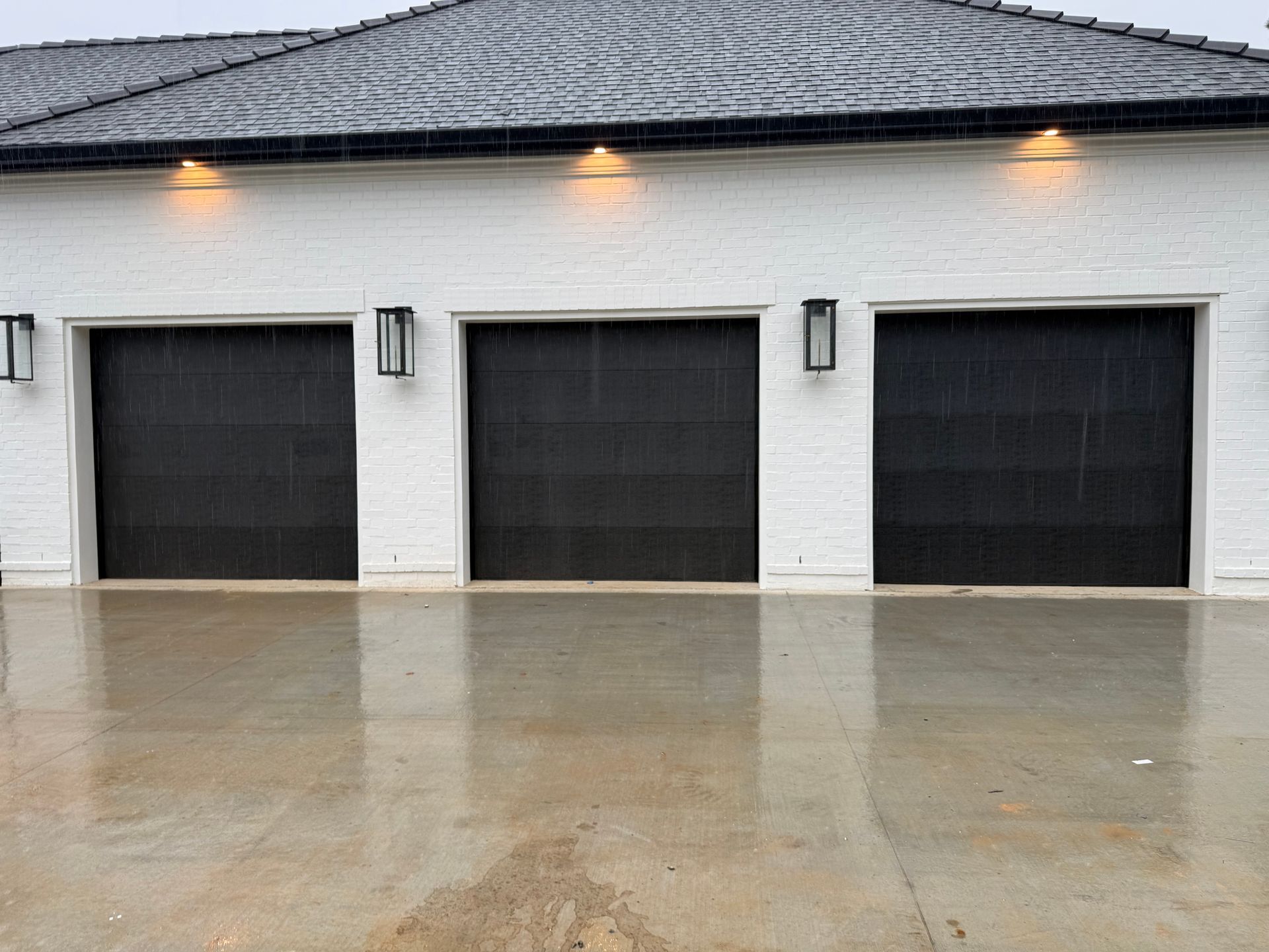 Three black garage doors on a white building, rain falling.