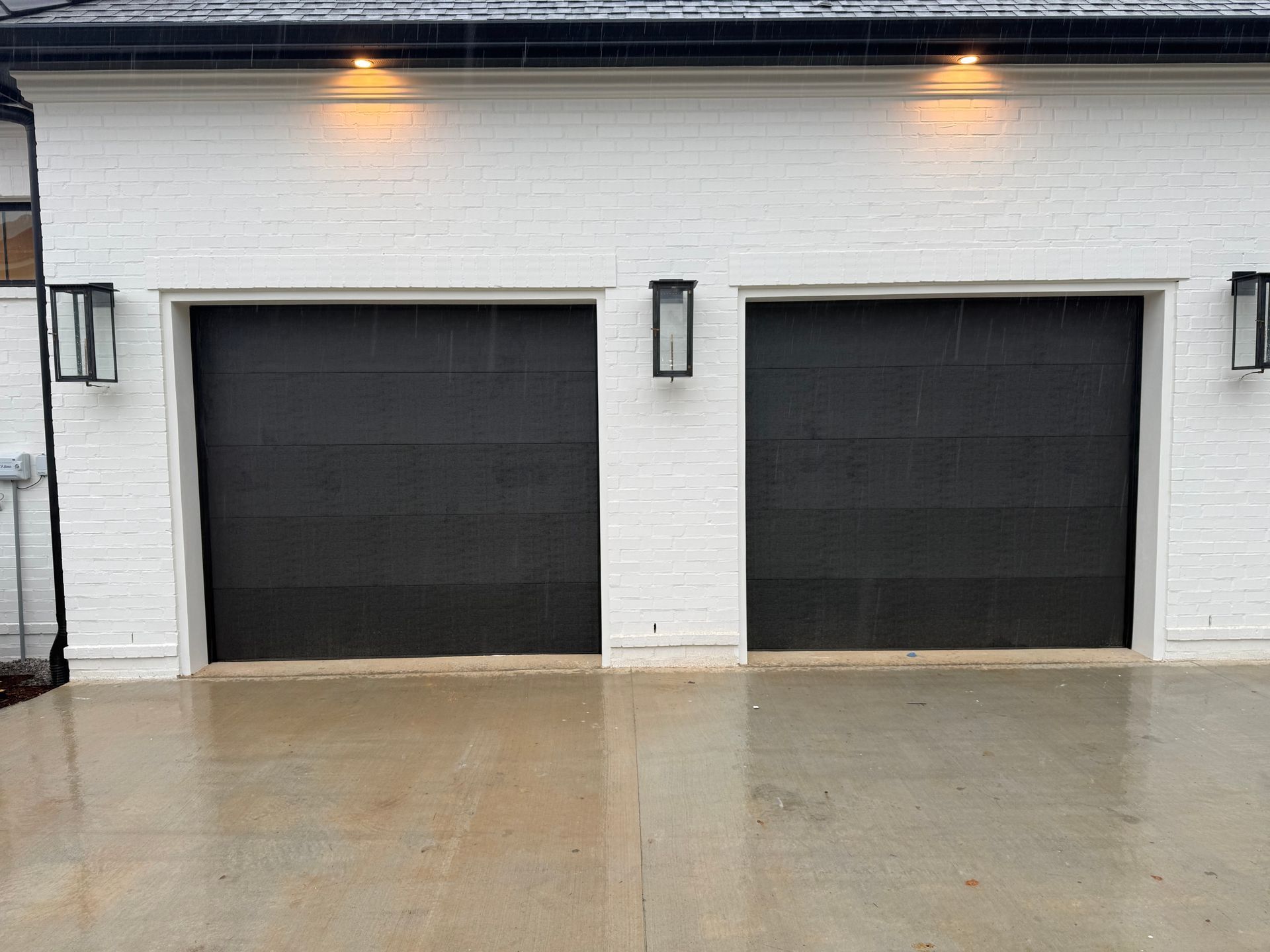 Two black garage doors on a white brick building with lights and a wet concrete surface.